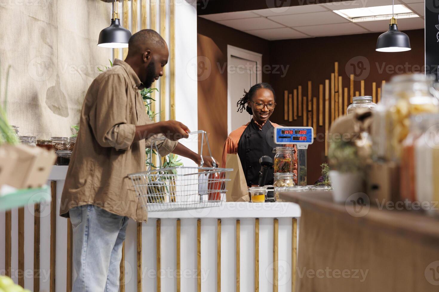 African American male customer unloading his shopping basket at the cashier counter. The friendly female vendor waits patiently with a smile and assists with the weighing of the products. photo