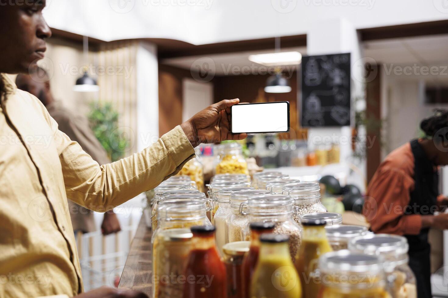 At eco friendly grocery shop African American male consumer holds a cell phone that displays an isolated white screen. Image shows black man with mobile device having a chromakey template. photo