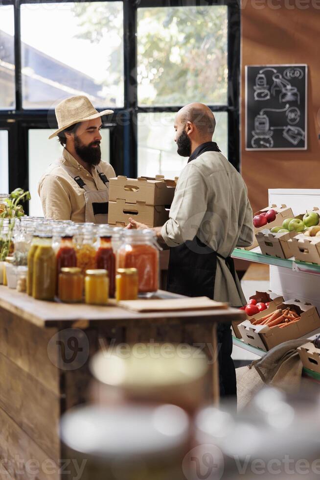 Caucasian man handing over boxes of freshly harvested items to middle eastern vendor with black apron in grocery store. Storekeeper receives new batch of locally grown produce from farmer. photo