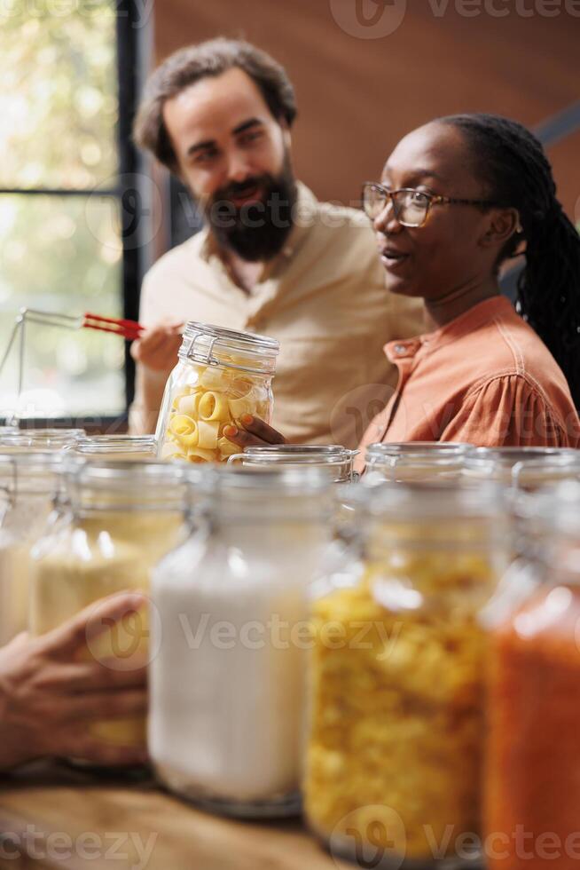 Portrait of african american woman holding a glass jar filled with pasta while standing next to caucasian man in local store. Multicultural couple doing sustainable shopping for healthy lifestyle. photo