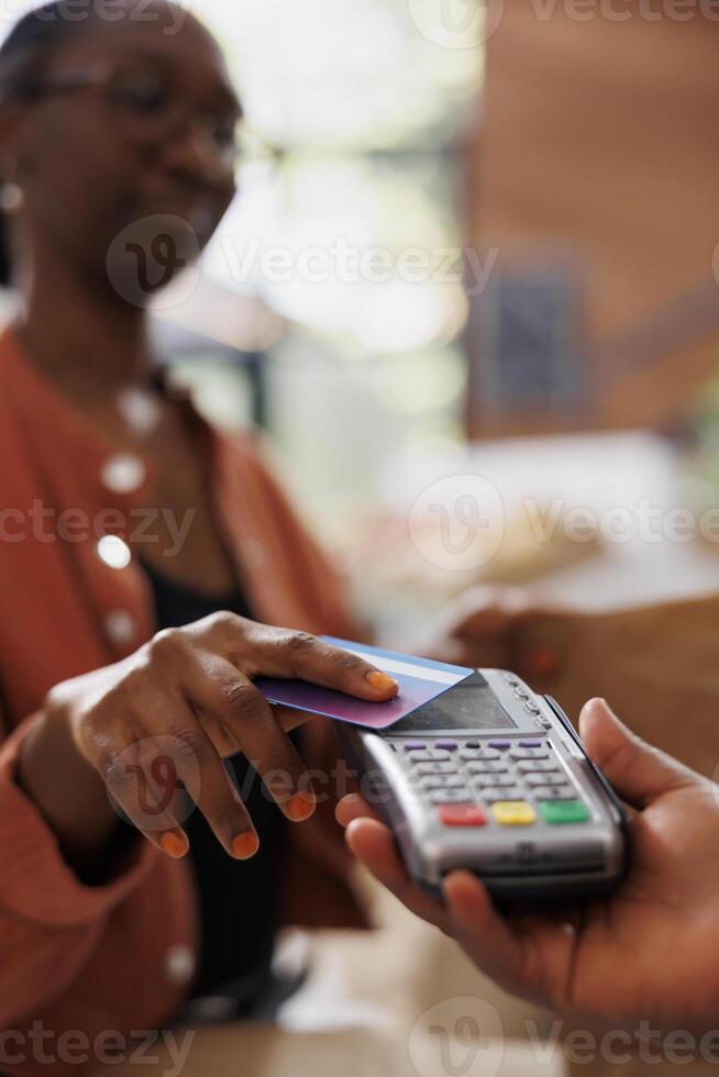 An African American individual making a contactless payment with a debit card to purchase fresh organic produce at a nearby market. Black woman paying with card at checkout counter. photo