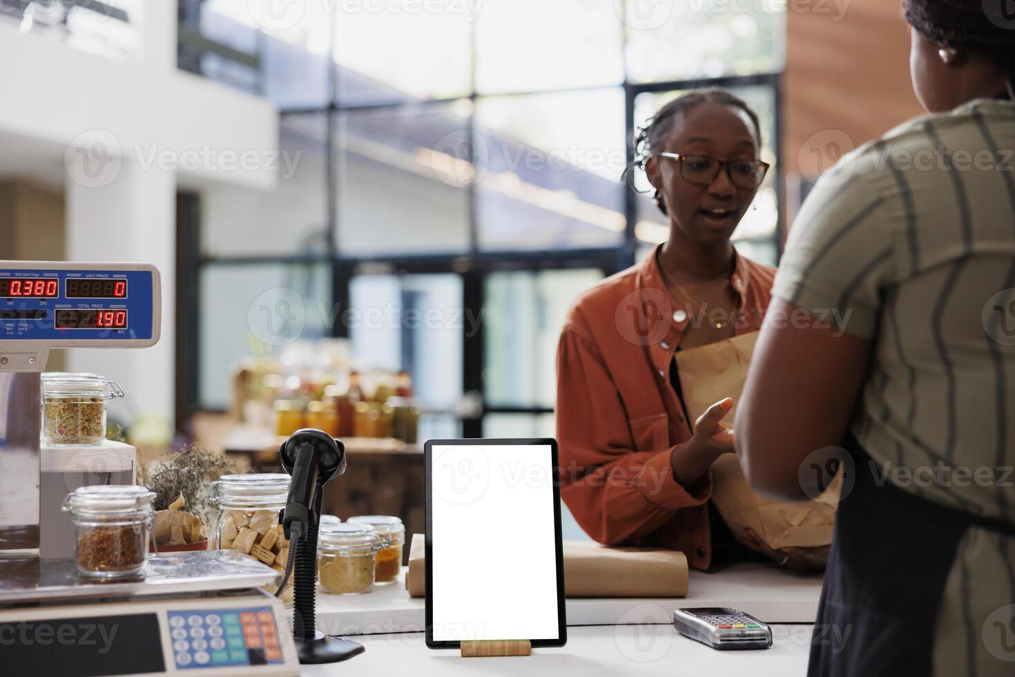 A lady wearing glasses converses with a vendor at the payment counter, while a digital device showcases a blank white screen featuring eco-friendly store advertisements. The tablet displays an empty chromakey mockup template photo