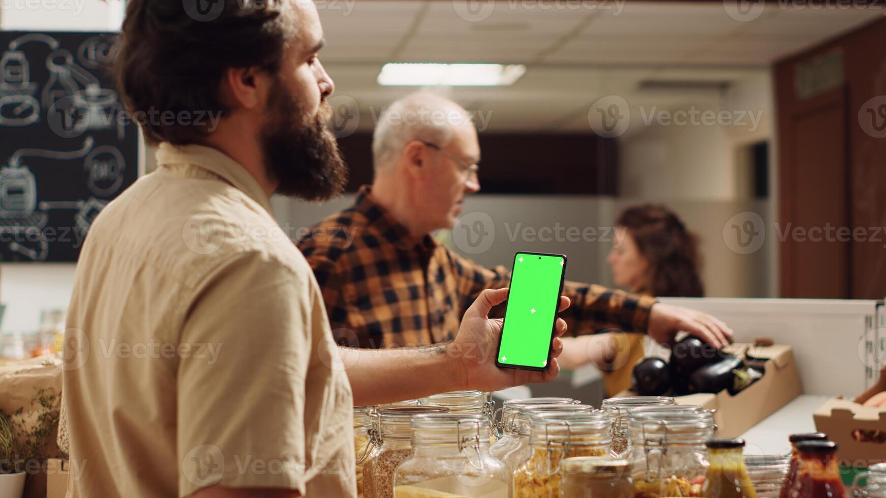 Man using green screen smartphone in zero waste supermarket to check ingredients for healthy recipe. Client in local grocery shop uses chroma key phone while shopping for organic veggies photo