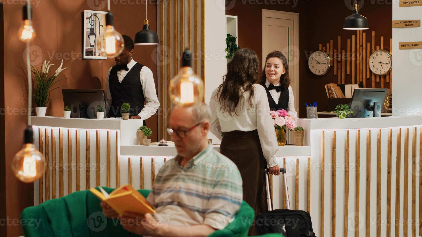 Arriving in modern hotel lobby, elderly woman arrives at front desk to do room check in. Senior tourist carrying baggage on vacation, staff members welcoming guest at reception. Tripod shot. photo