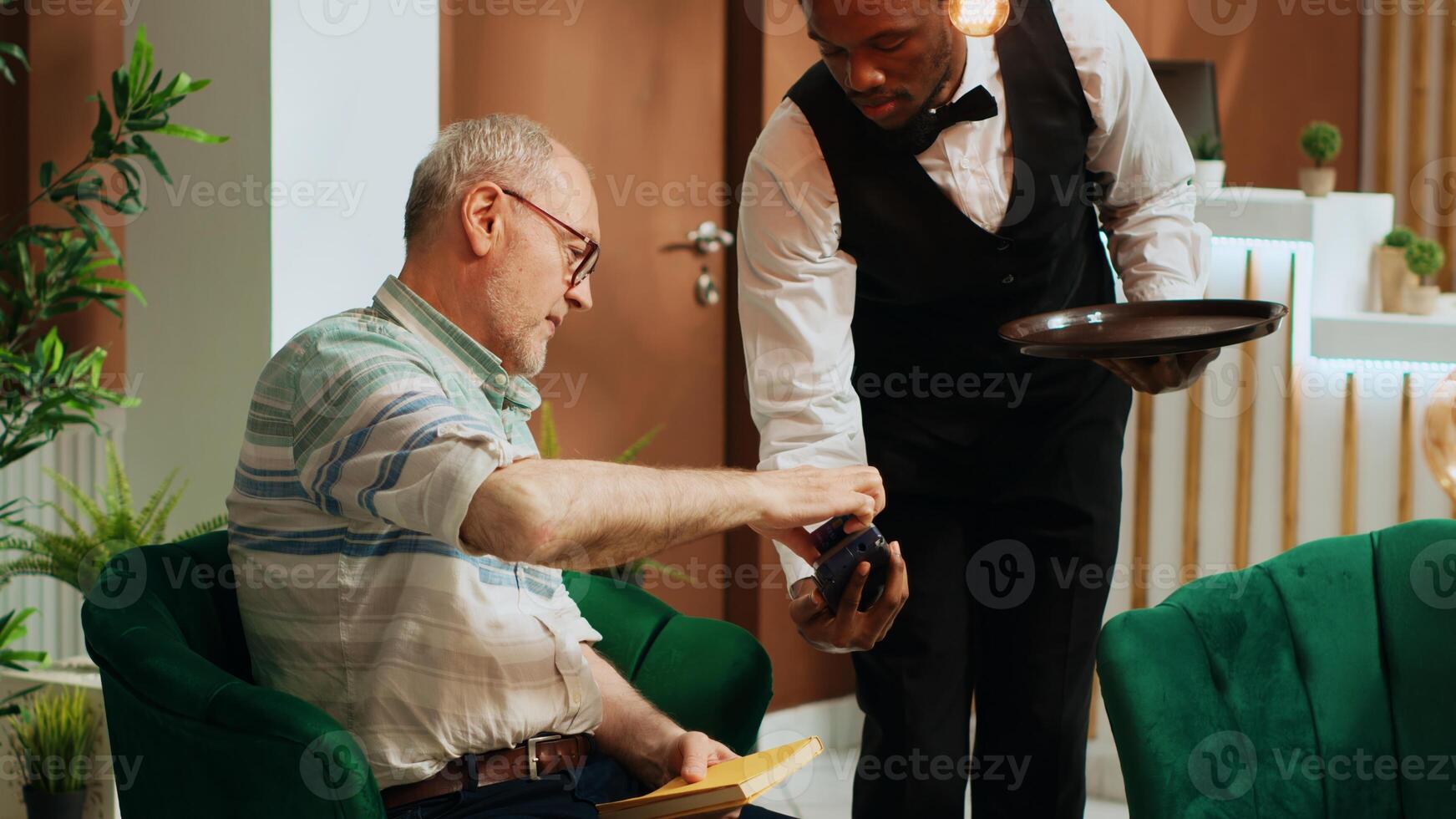 Elderly person paying for coffee cup via credit card in hotel lobby, ordering refreshment from bar and making pos transaction. Bellhop serving guest with beverage while he is waiting. photo
