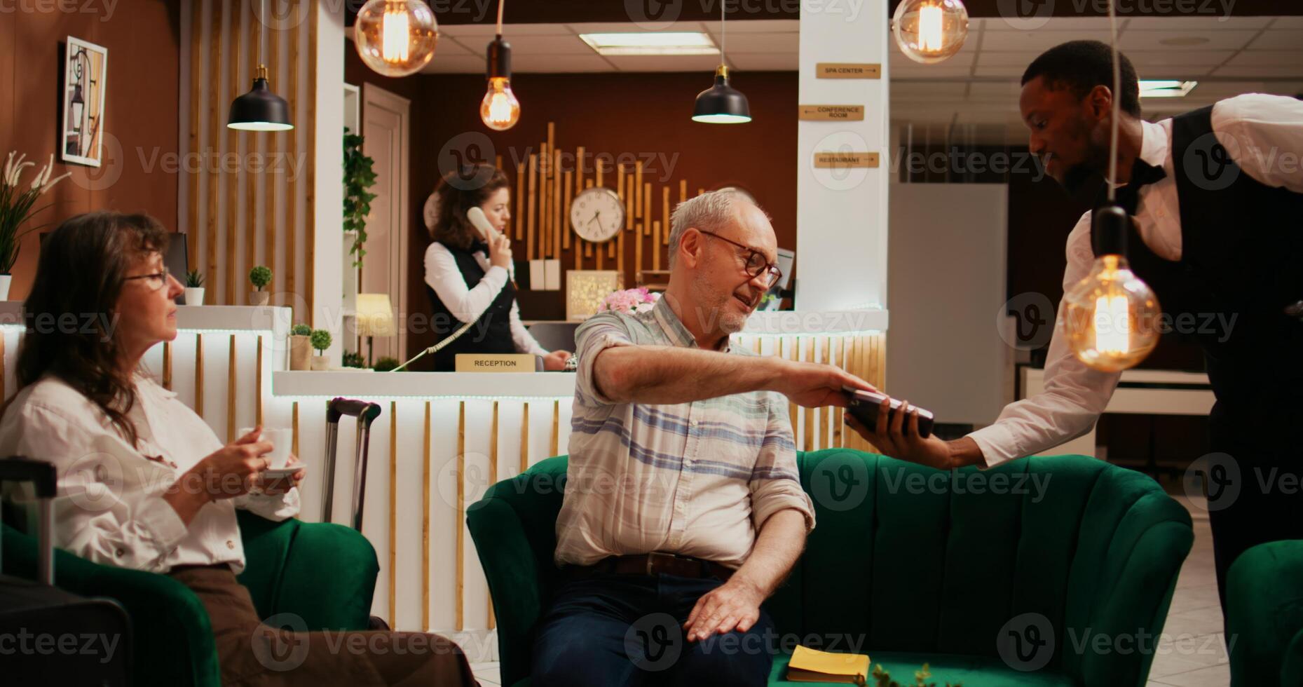 Retired couple paying for coffee cup from hotel bar, taking out credit card for pos terminal transaction. Husband and wife relaxing together in lounge area at resort, paying for services. photo