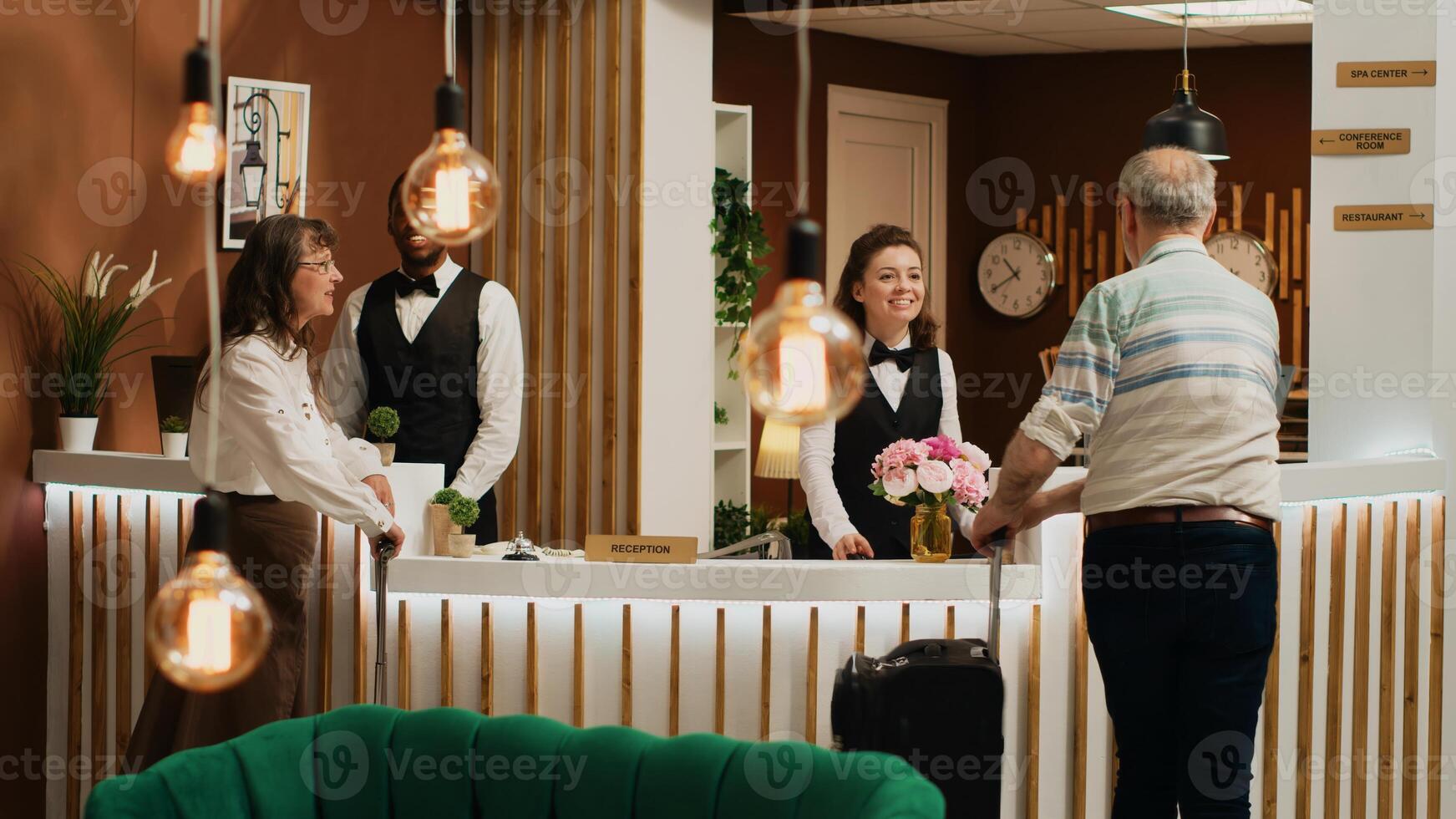 Senior couple arrive in hotel lobby, talking to front desk employee before check in process. Tourists travelling on vacation for leisure activities, hotel concierge services. Tripod shot. photo