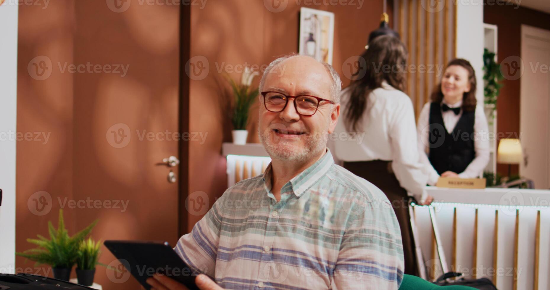 Senior person using modern tablet in hotel lobby, relaxing in lounge area before starting retirement holiday trip. Browsing online webpages to find recreational activities, leisure senior travel. photo