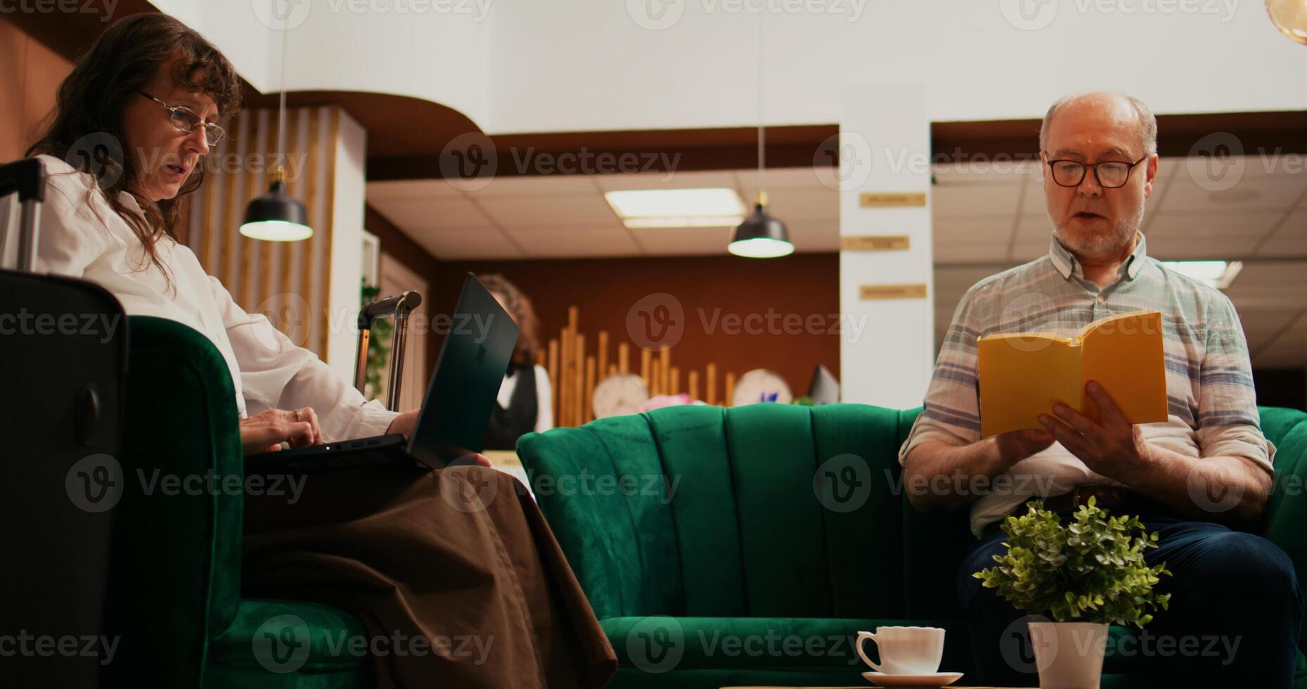 Senior couple passing time at hotel, wife creating trip itinerary on laptop while husband reading book in lounge area. Retired people travelling on international vacation, waiting for check in. photo