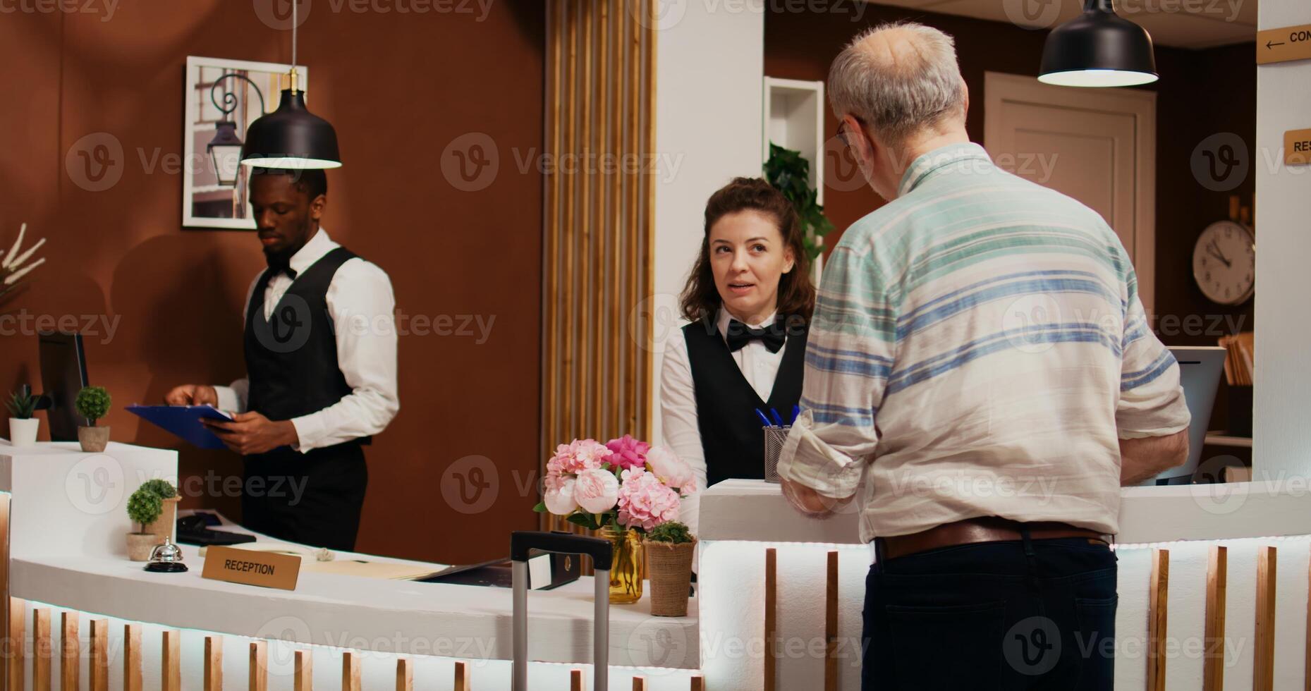 Elderly person arriving at hotel front desk with trolley bags, asking for assistance to check in. Receptionist ensuring pleasant stay for old man at all inclusive holiday retreat, tourism industry. photo