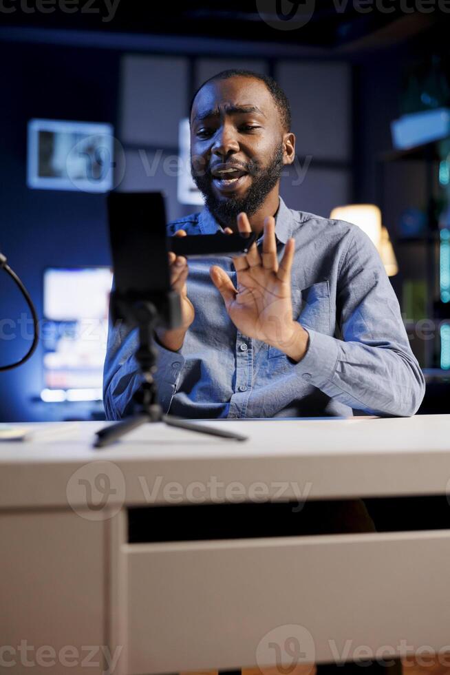 Influencer in home studio using smartphone on holder to record live broadcast, reviewing mobile phone. Online show host filming electronics haul, presenting newly launched cellphone for viewers photo