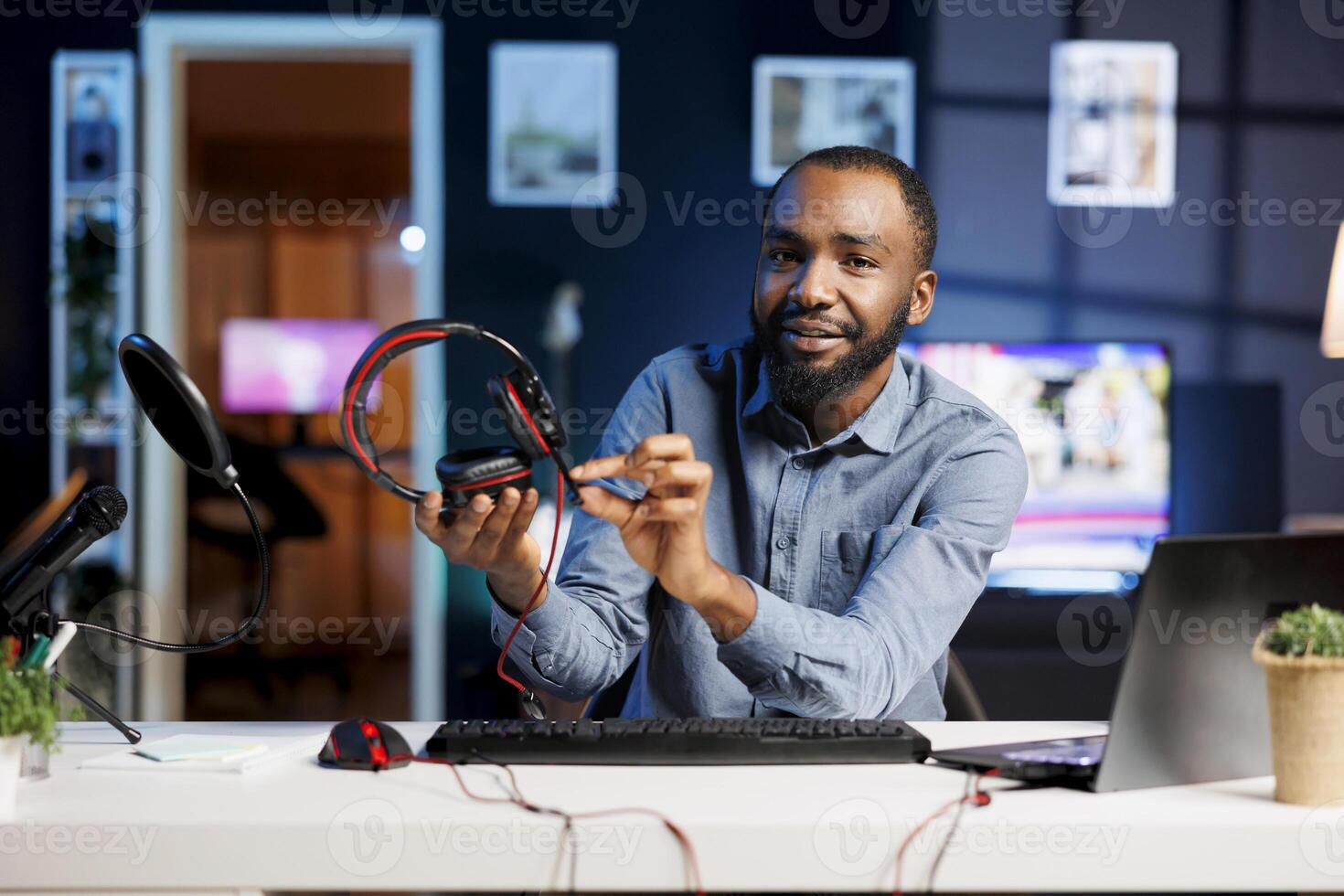 African american man recording for internet channel in dimly lit home studio, presenting gaming headphones to subscribers. Media star showing viewers audio listening device photo