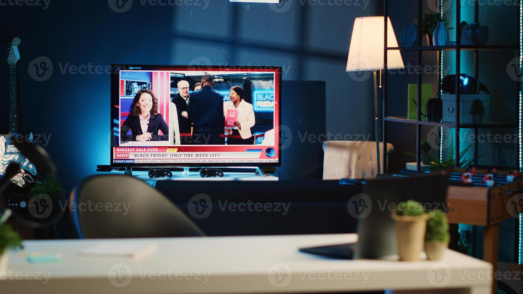 Apartment illuminated by RGB lights with TV broadcast showing Black Friday news story. Blurry foreground of work desk with laptop and mini house plants in dimly lit empty home studio interior photo