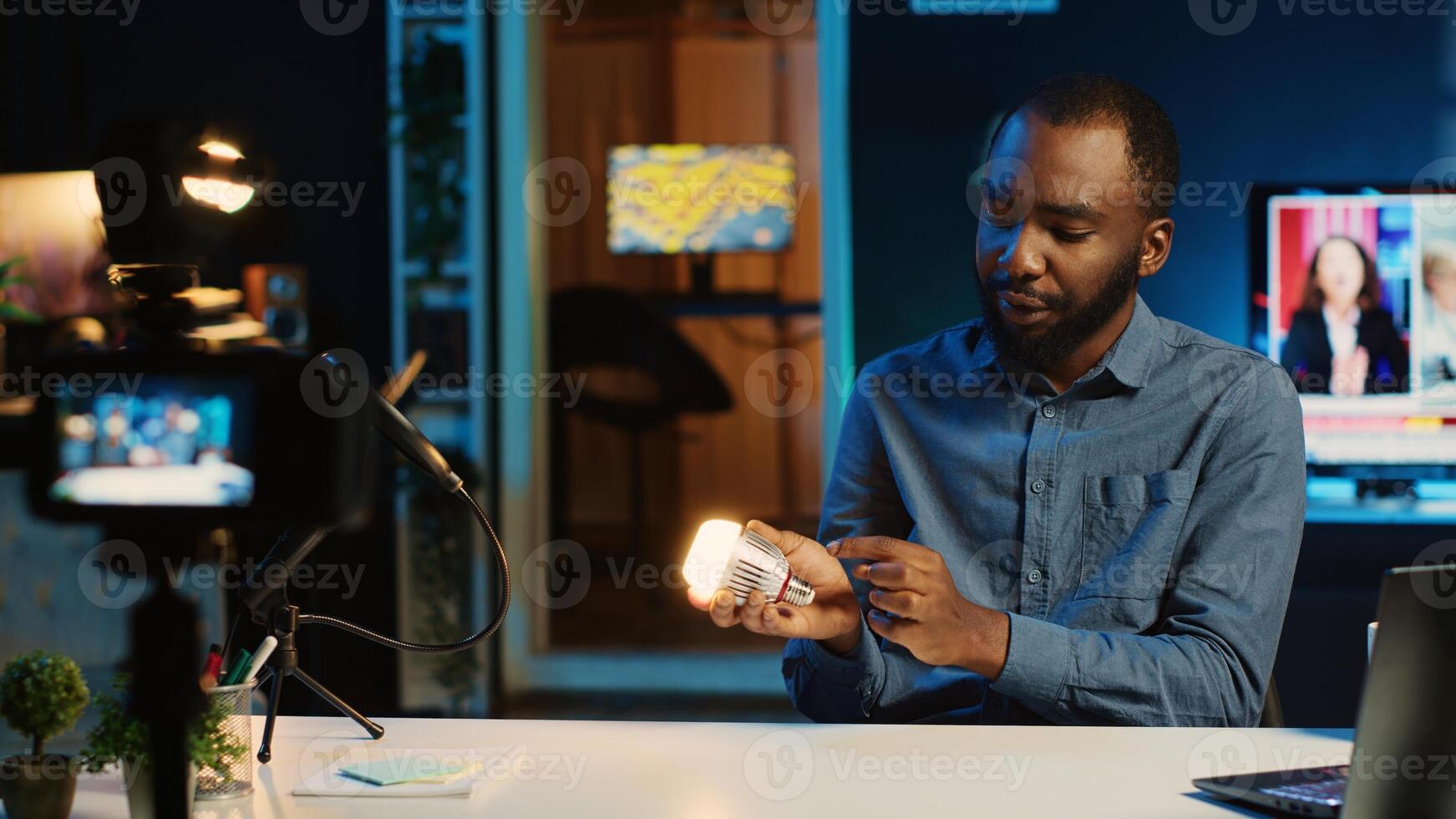 BIPOC content creator in dimly lit living room uses camera to film smart light bulb review for online streaming platforms. Media star hosts internet show, unboxing home automation product photo