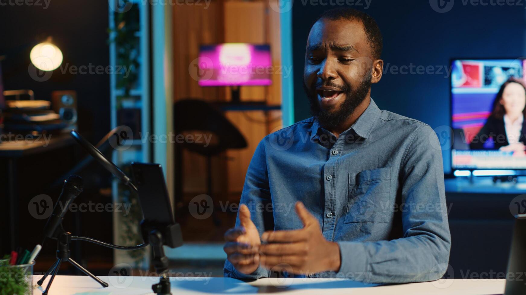 Content creator in dimly lit home studio using cellphone camera to film for online streaming platforms. Influencer greets audience using mobile phone to record footage photo