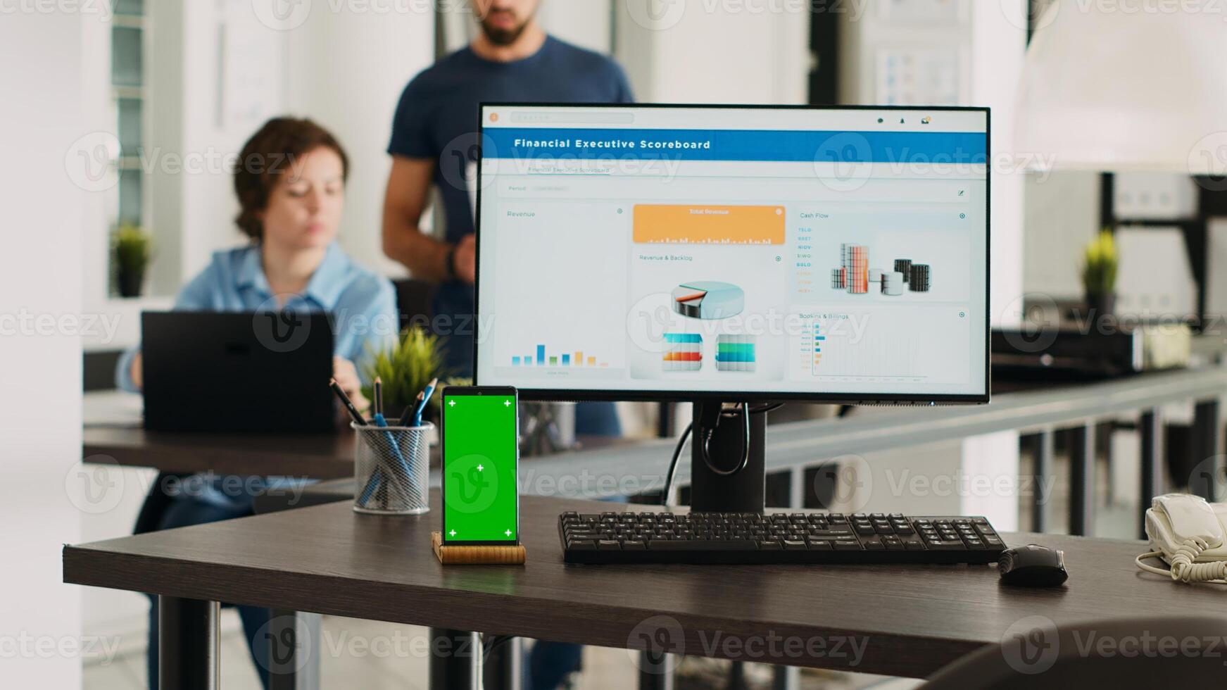 Persons conducting business tasks at agency workspace with cellphone displaying greenscreen at empty desk. Isolated copyspace mockup layout appears on workstation with mobile device. photo