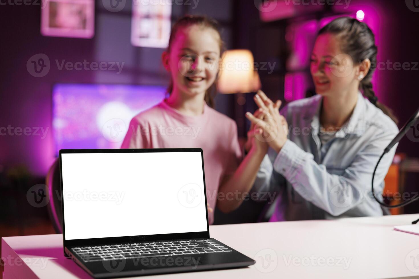 Cheerful girl next to her mother presents mockup laptop in pink neon lit living room used as vlogging studio. Young media star helped by parent to showcase isolated screen notebook device photo