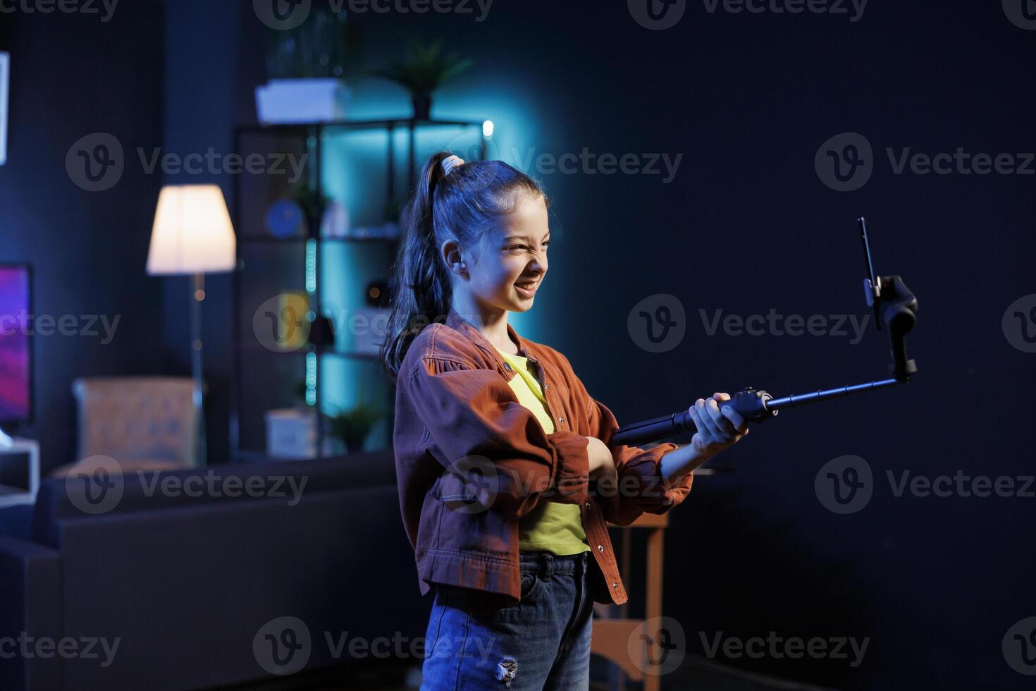 Cute kid doing content creation for children, utilizing cellphone on selfie stick to produce dance videos for online streaming platforms. Excited child creating viral choreography with phone photo