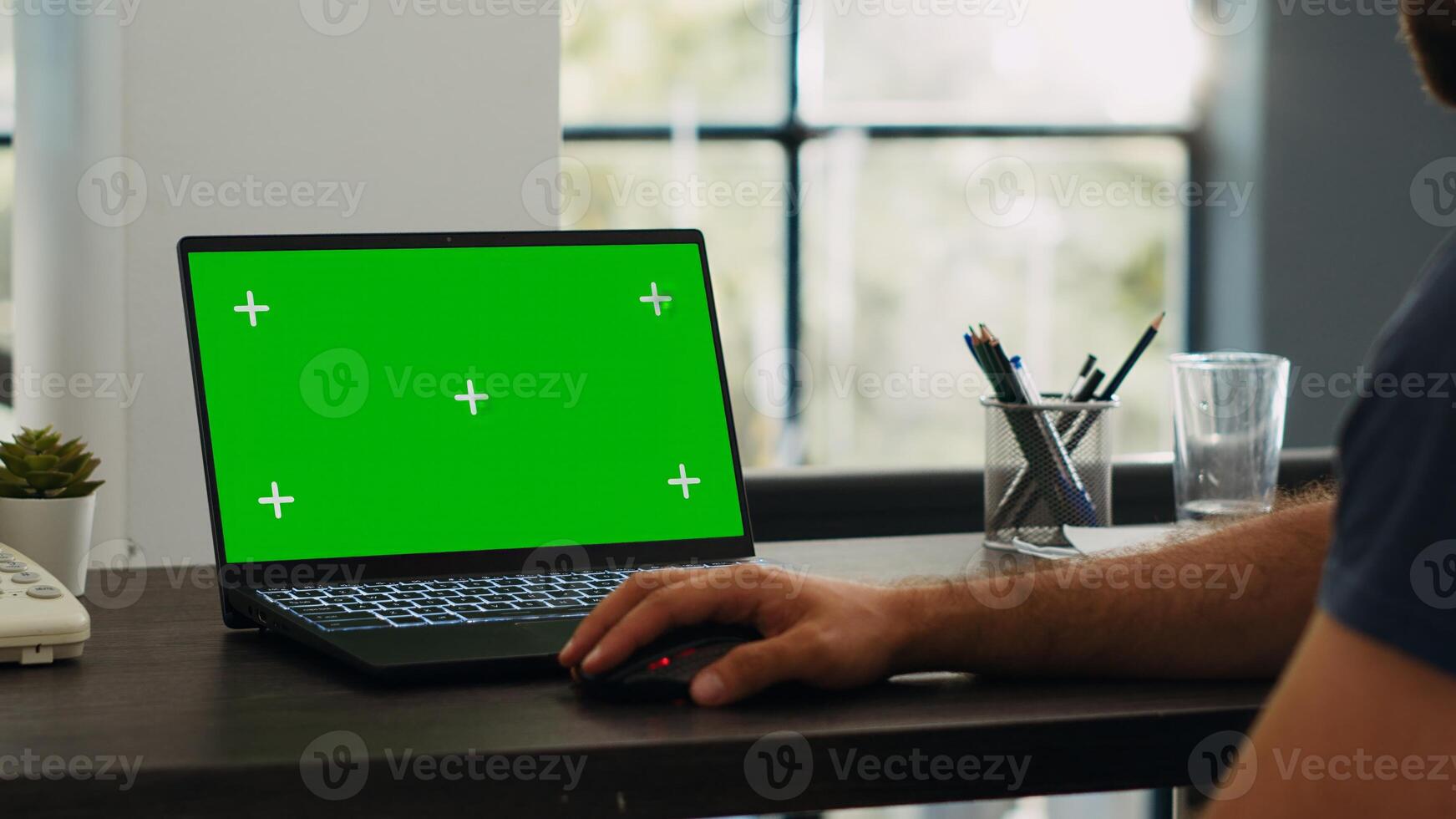 Male employee looks at greenscreen chromakey display while he sits at creative agency workstation, working on e commerce operations. Professional analyst checks blank mockup layout on laptop. photo