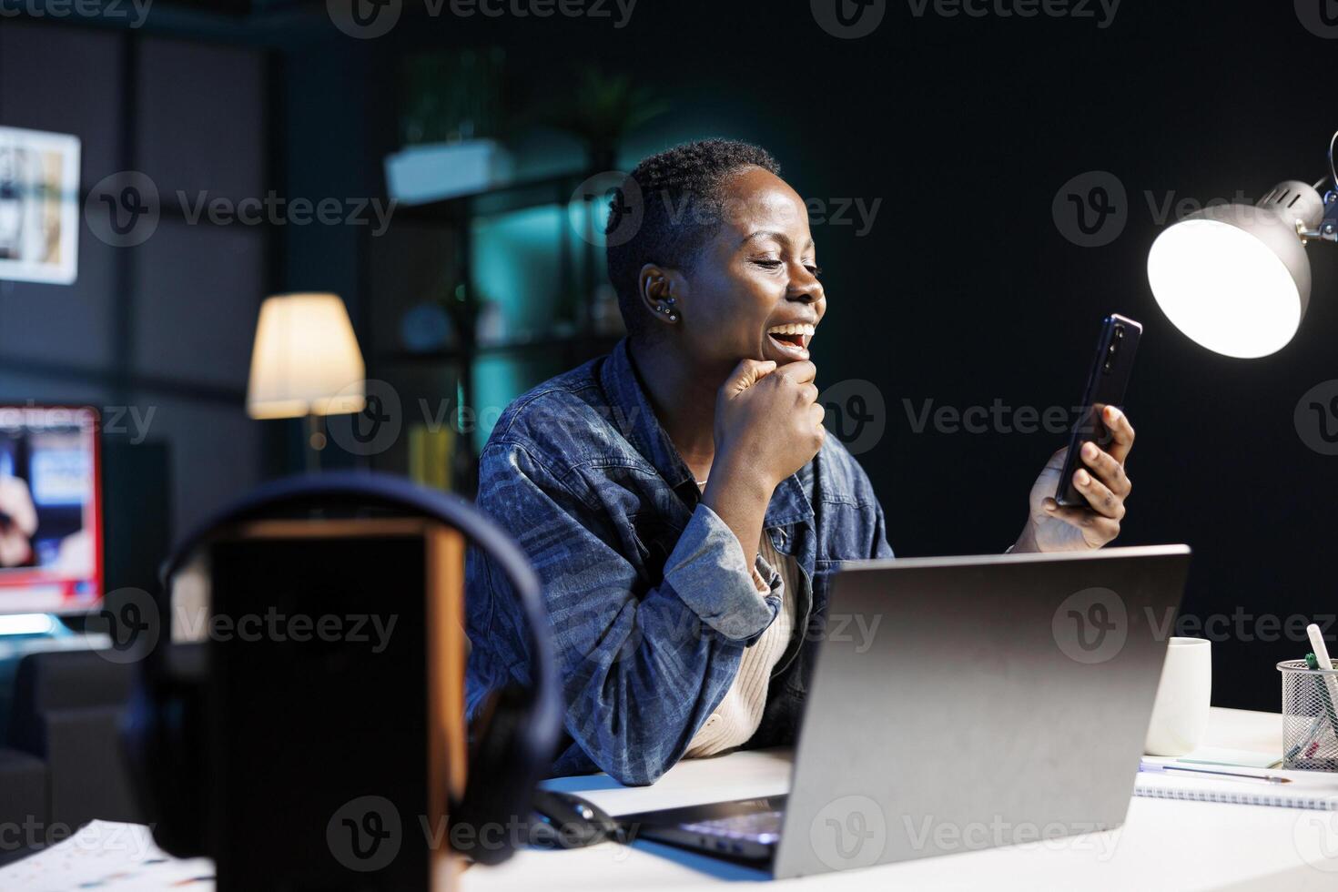 Excited black woman working remotely, using mobile device for calls. Cheerful female blogger uses a laptop and cellphone to browse the internet, check emails, and stay connected. photo