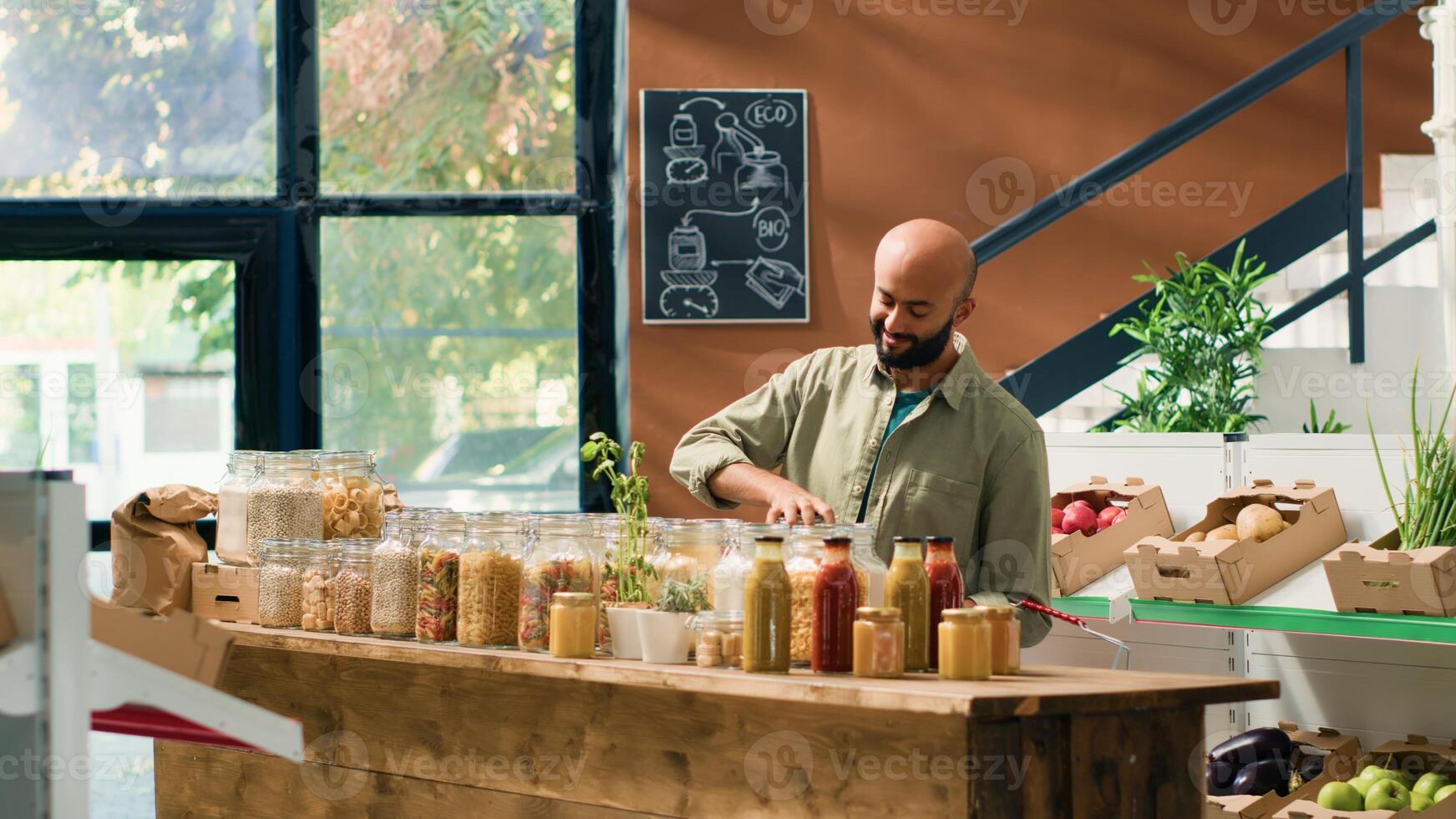 Middle eastern man in ecological food store examining organic homegrown fruits and vegetables, opening jars with sustainable pasta or spices. Customer admires fresh bio merchandise. photo