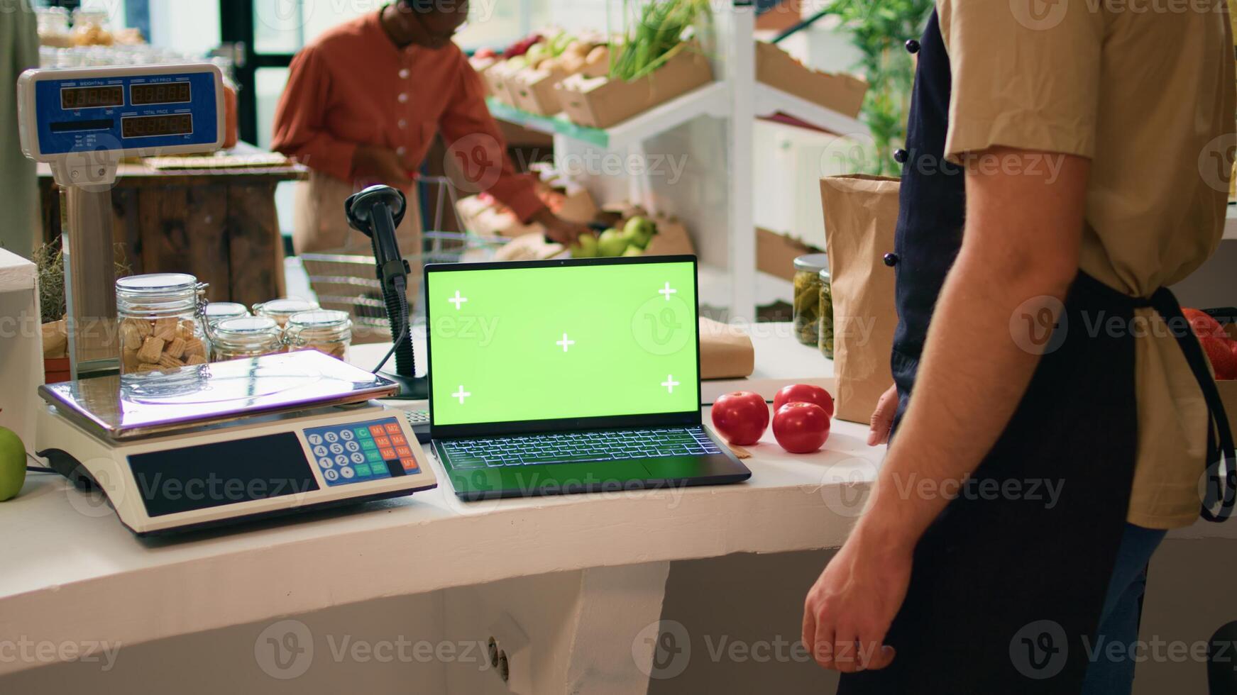 Shop owner works with greenscreen on laptop, sitting at register counter and waiting to serve customers in local zero waste supermarket. Vendor using blank display with chromakey mockup. photo