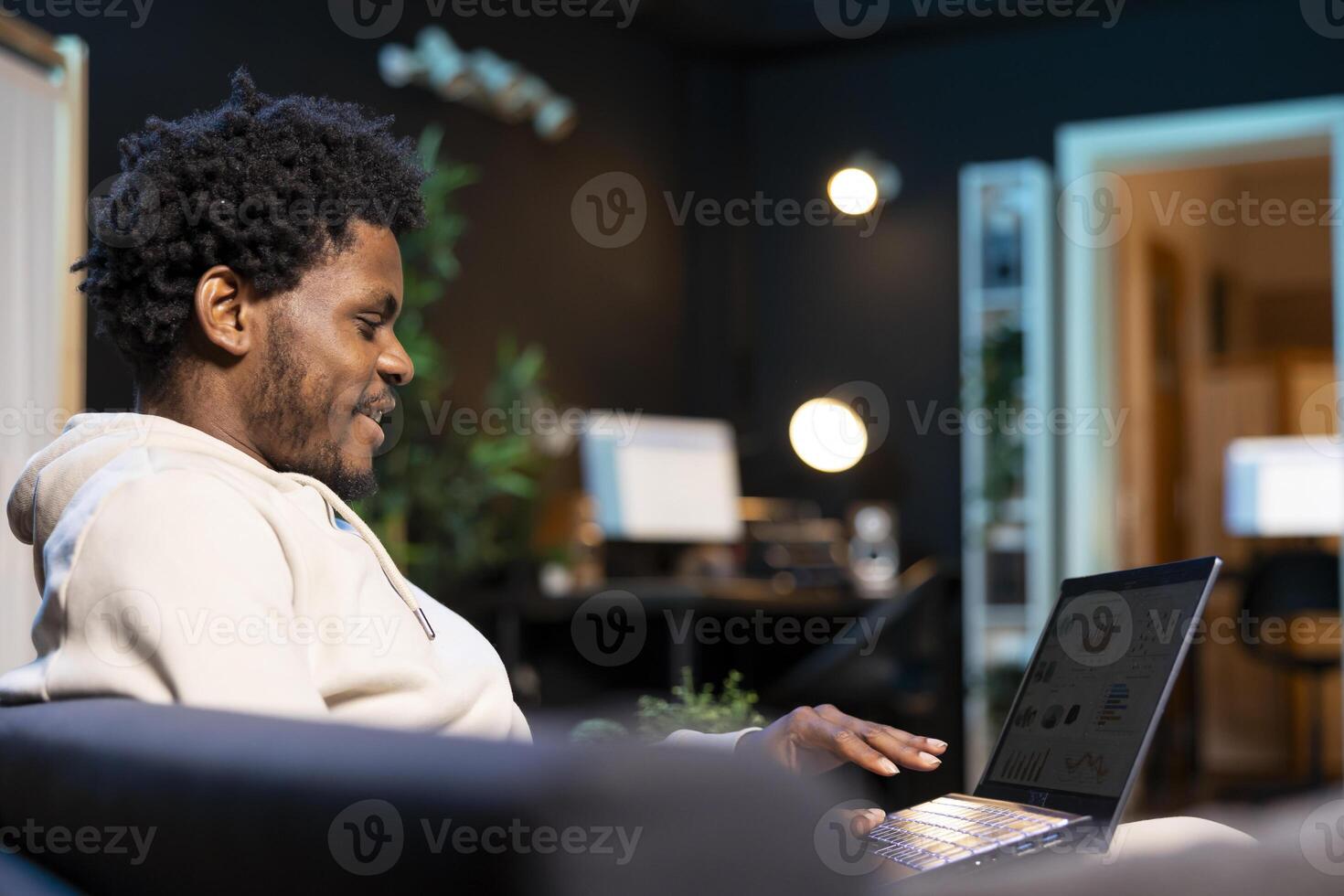 Smiling teleworker analyzing financial graphs in stylish apartment, happy about finishing work soon. Cheerful african american employee remotely working, looking at business charts on laptop photo