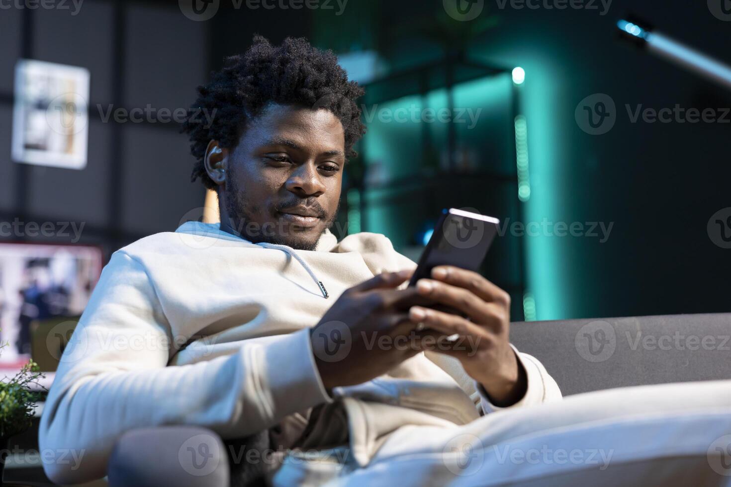 Smiling man impatiently texting friend, planning meeting hour together. Person sitting on cozy sofa in modern stylish apartment typing on cellphone, excited to meet up with mate photo