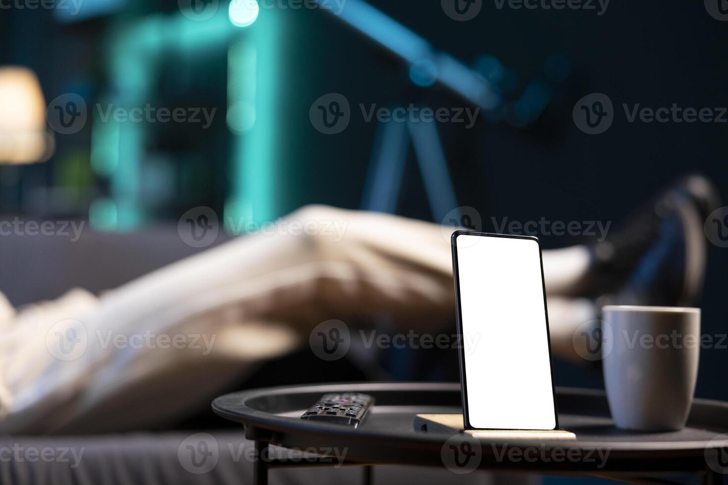 Focus on mockup device with man sleeping on couch in blurry background in neon lit apartment. Close up of isolated screen smartphone on coffee table in front of person snoozing photo