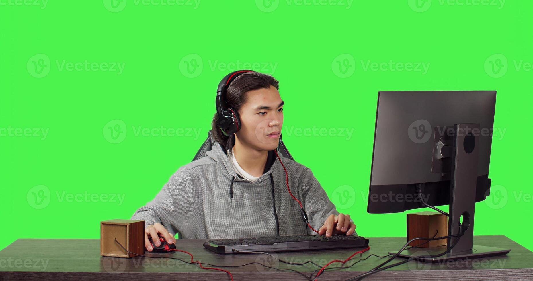 Asian person gaming on pc at workstation, playing online videogames with friends. Young man having fun with multiplayer contest, speaking to people on headphones over greenscreen. photo