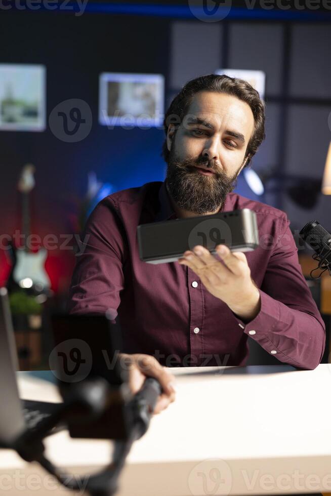 Media star doing technology review of wireless portable speaker for, filming with phone on selfie stick. Influencer showcasing music playing device, capturing footage with cellphone camera photo