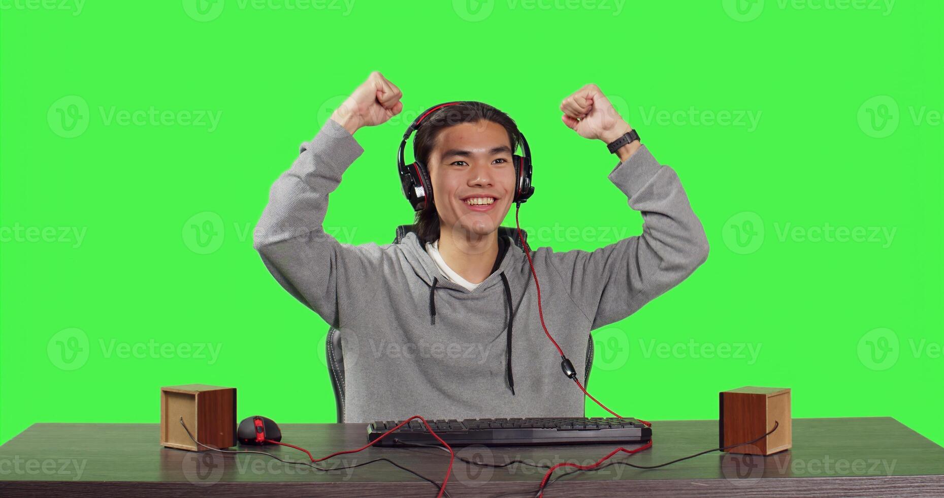 POV of asian adult enjoying computer games at desk while sitting over full body greenscreen backdrop. Gamer has fun with friends online in gaming web challenge, using headphones at desktop. photo