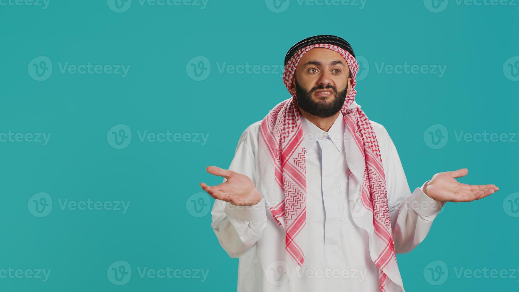 Muslim person being confused and unsure on camera, creating i dont know symbol and acting clueless over blue background. Young man in arab clothing being uncertain and shrugging. photo