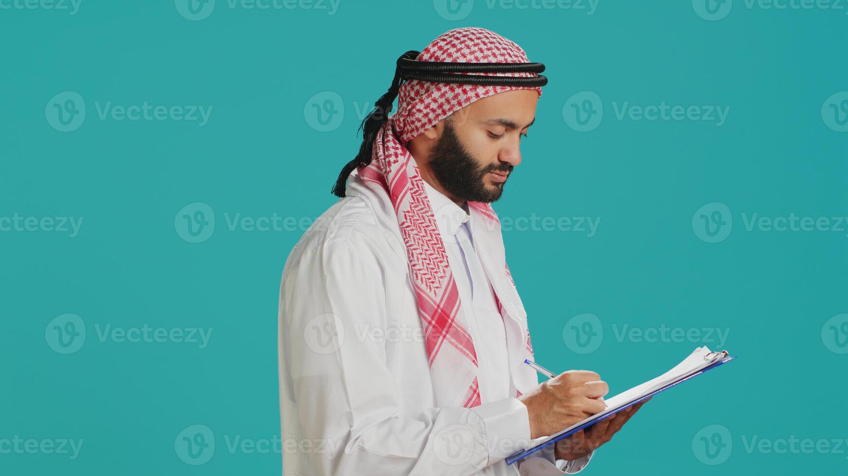 Arab man writing letter on papers standing over blue background, creating new schedule and taking notes. Young adult in muslim clothing looking at checklist for paperwork. photo