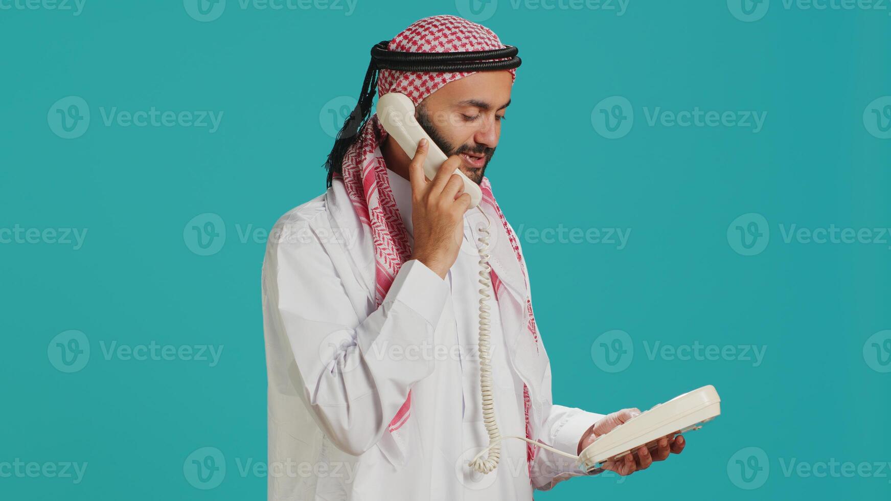 Arab guy talks on landline phone call in studio, answering call on retro telephone with cord for remote conversation. Young adult communicating with people on office phone line. photo
