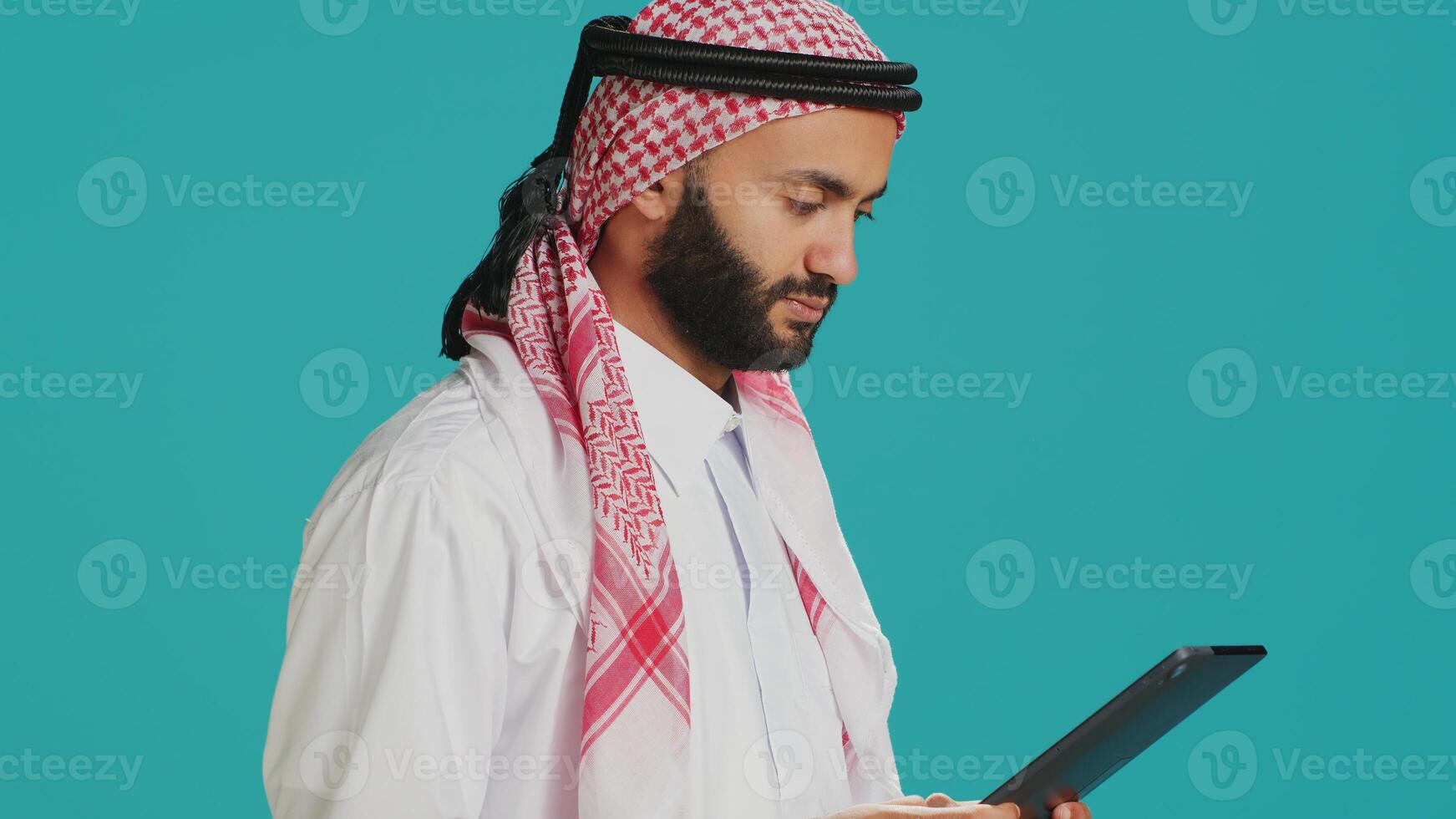 Middle eastern adult browsing device in studio, navigating websites online on digital gadget. Muslim person in traditional clothes using tablet to check internet pages, texting on social media. photo