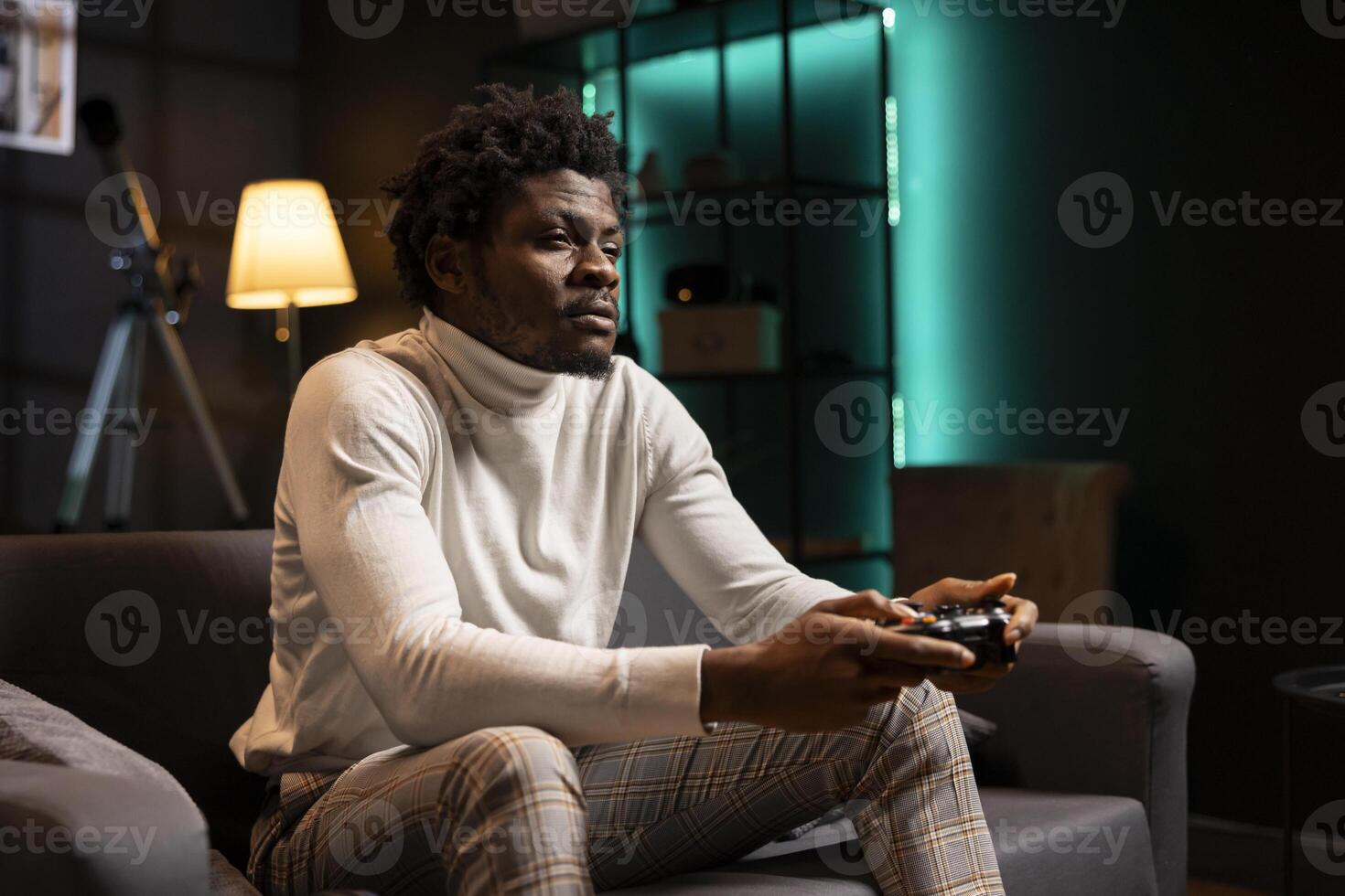 BIPOC player at home gaming and enjoying leisure time. Man sitting on couch in cyan neon illuminated living room, playing videogame on console, relaxing after hard day at work photo