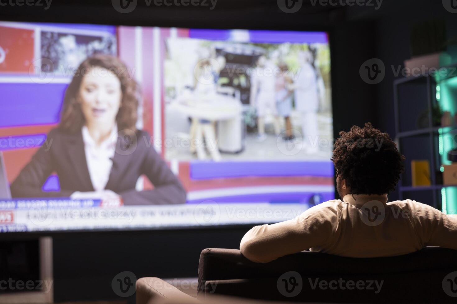 Man sitting on couch in front of ultrawide smart TV displaying news program broadcasting. Person watching big television screen showing VOD channel with newscast coverage photo
