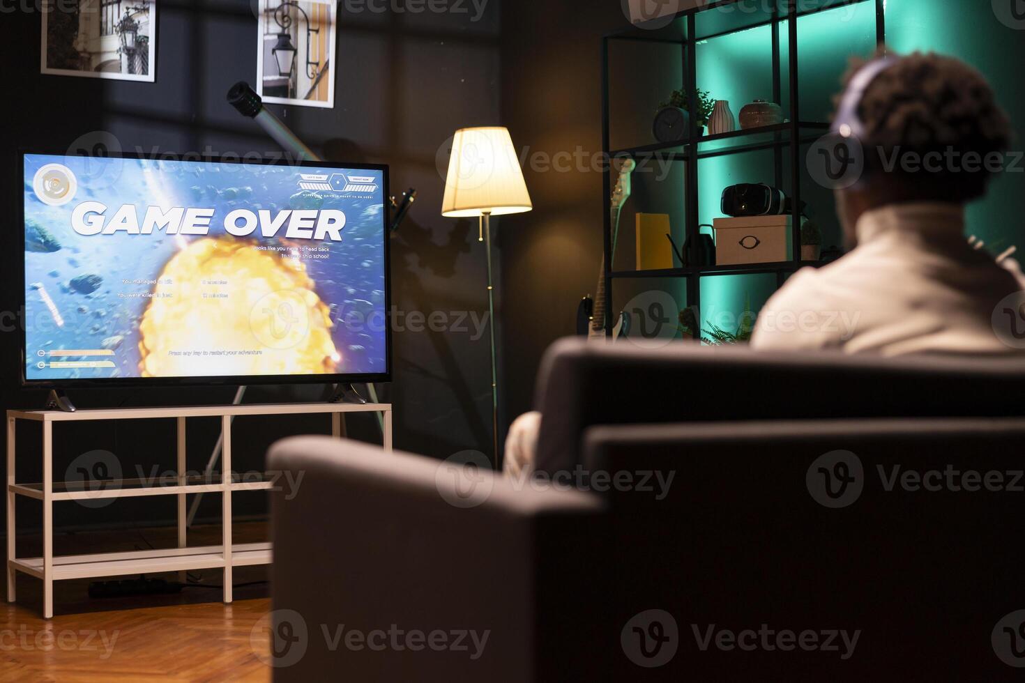 Gamer wearing headphones frustrated by seeing game over message on smart TV while playing arcade space shooter videogame. Man in home theatre spending time on gaming console, disappointed after losing photo