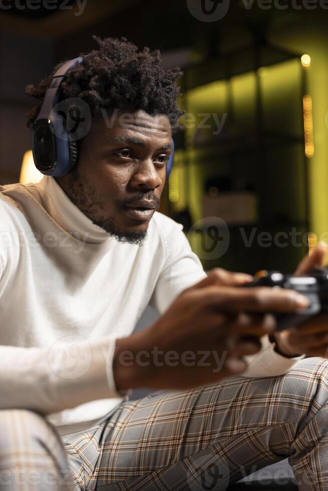 African american man playing videogame in yellow neon lit apartment, holding controller. Gamer participating in PvP online multiplayer game using console system and headphones for immersive experience photo