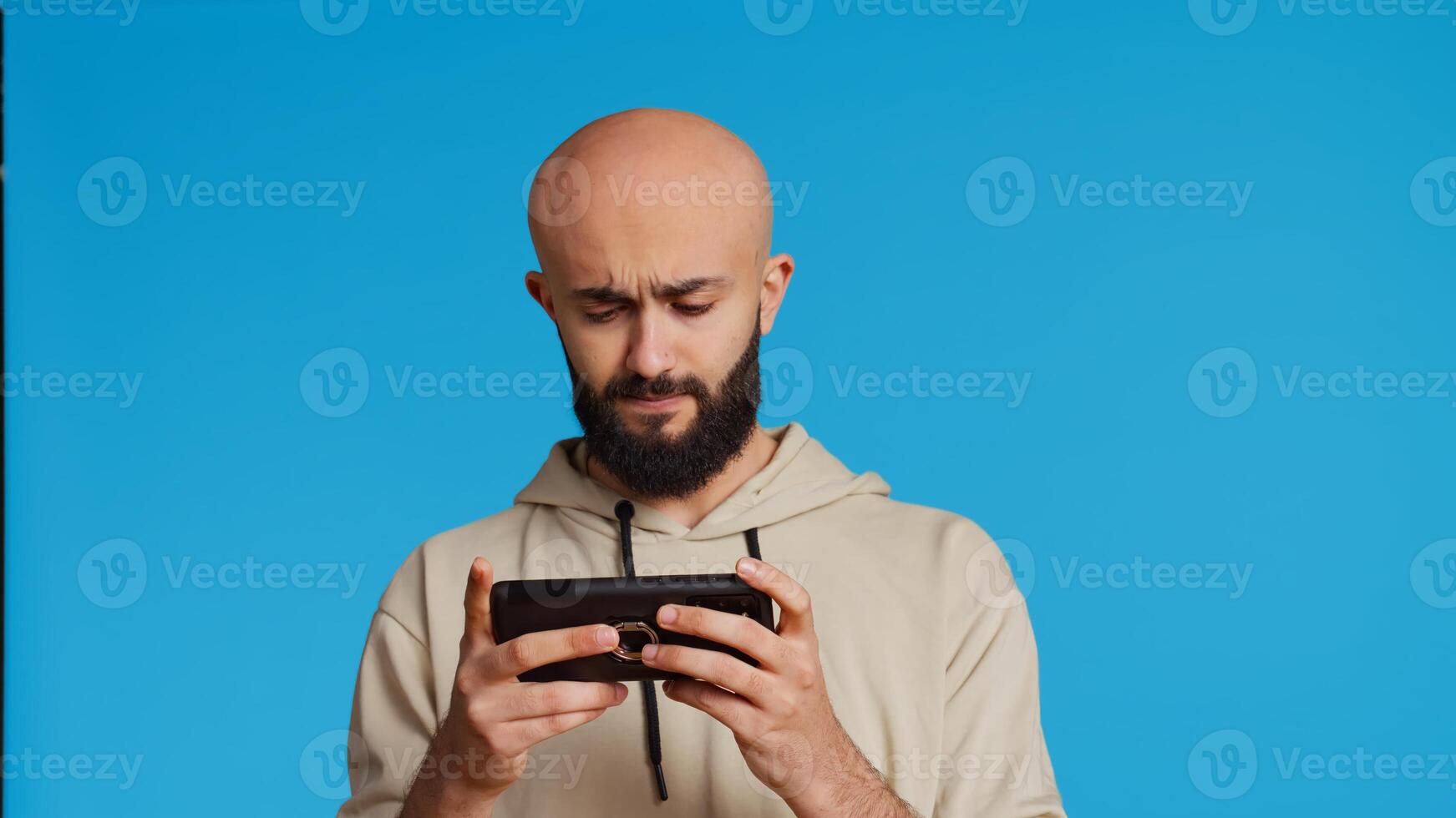 Middle eastern person playing mobile games on app, standing over blue background and enjoying online tournament using mobile phone. Man having fun with gaming contest. Camera 2. photo