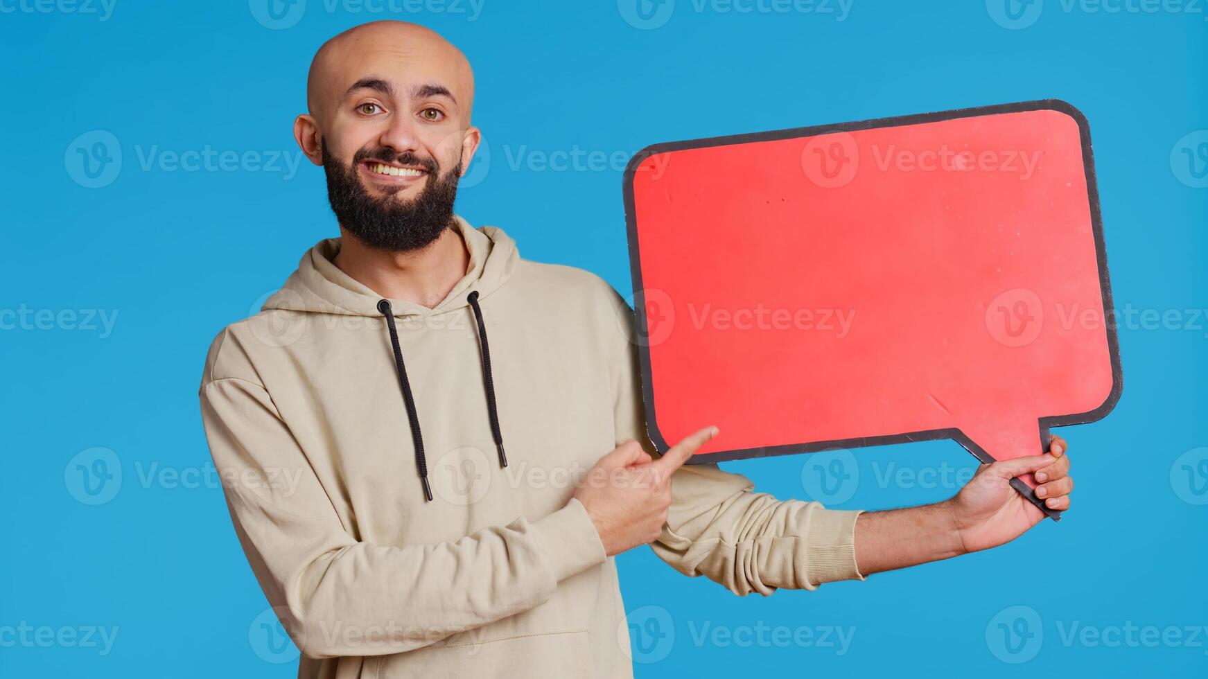 Arab person holding red speech bubble icon in studio, creating web advertisement for new product commercial. Middle eastern man shows isolated cardboard sign for ad. Camera 2. Handheld shot. photo