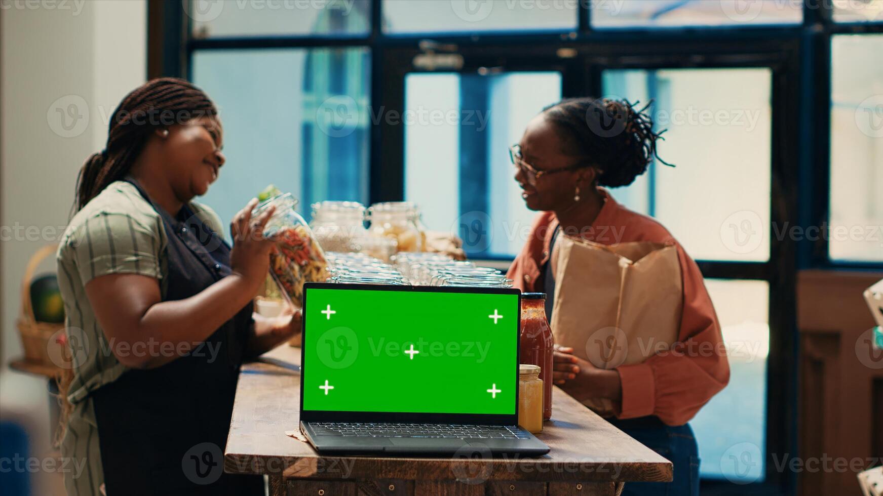 Vendor recommending natural bulk items in jars, talking to client next to laptop showing isolated greenscreen display in local supermarket. Farmers daughter presents homemade grains and spices. photo