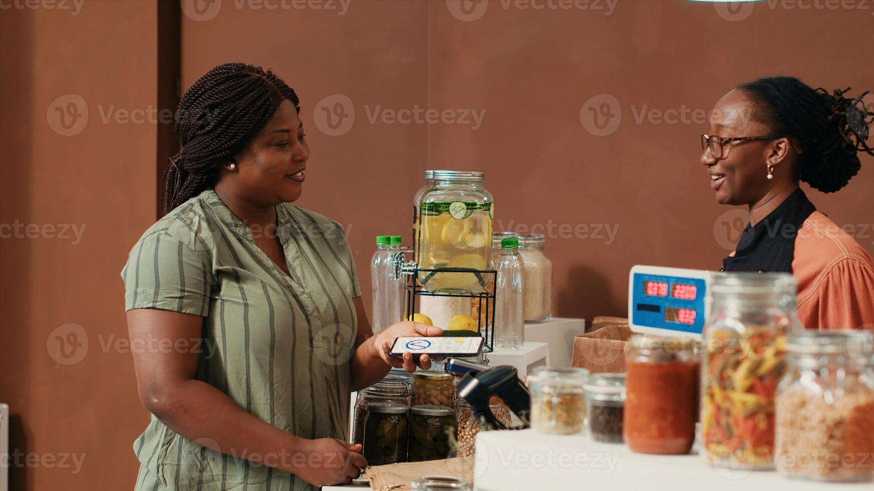 African american woman paying with mobile nfc at pos, buying fresh eco friendly produce from local farm. Young vendor helping client to weight merchandise and bulk products on scale. photo