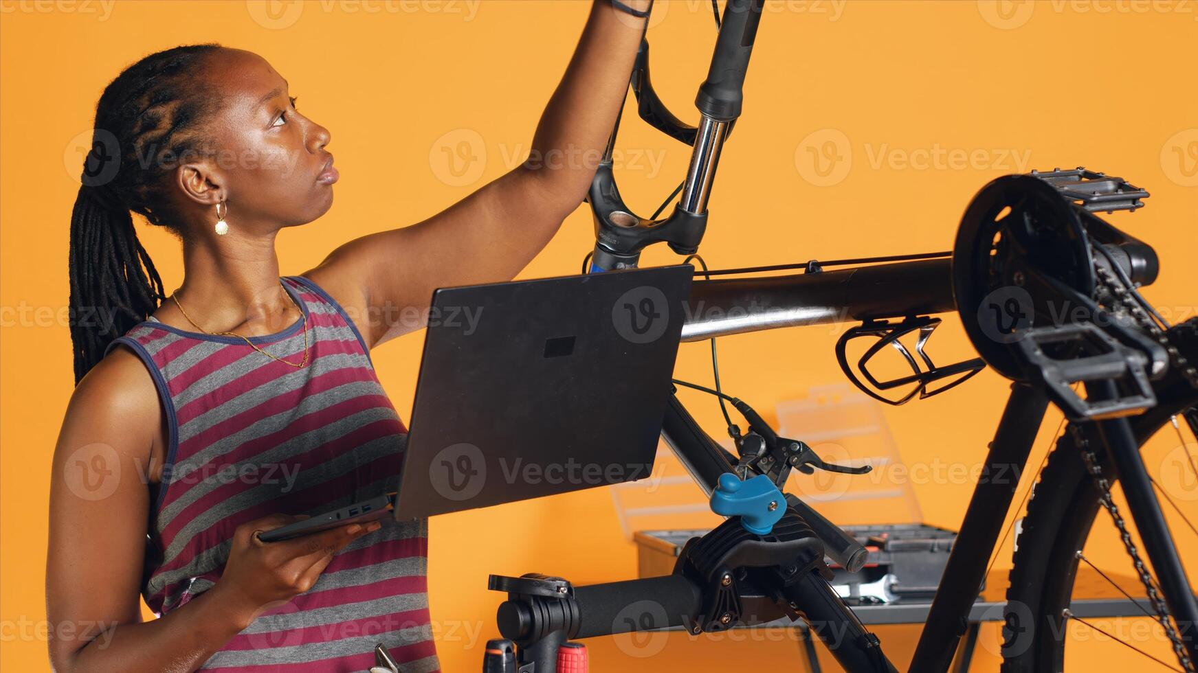 African american engineer doing bike servicing in studio background repair shop, looking on laptop maintenance list. Professional checking bicycle parts that need fixing, camera A photo