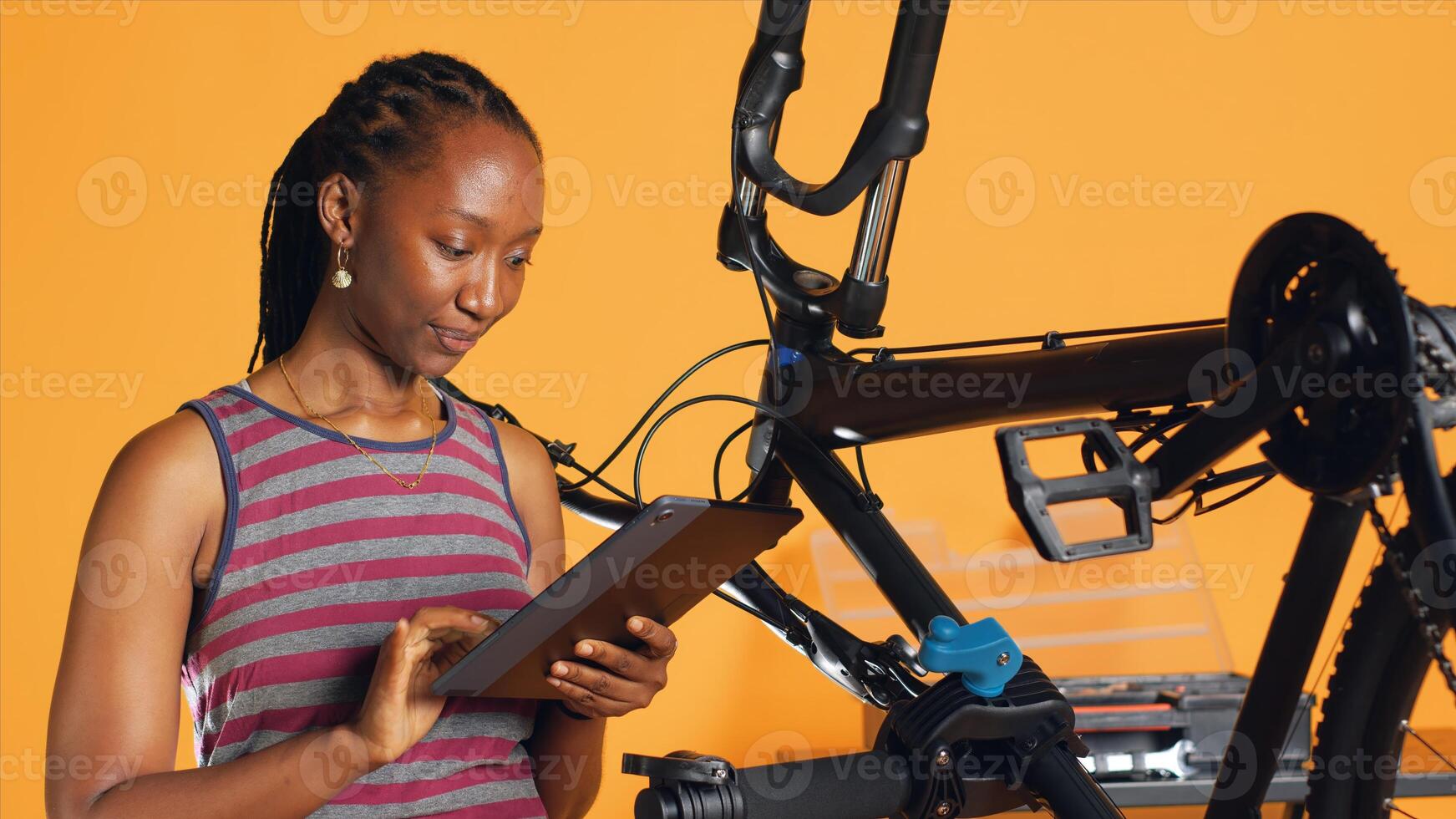 African american woman doing bike servicing in studio background repair shop, looking on tablet maintenance list. Technician checking bicycle components that need repairing, camera A photo