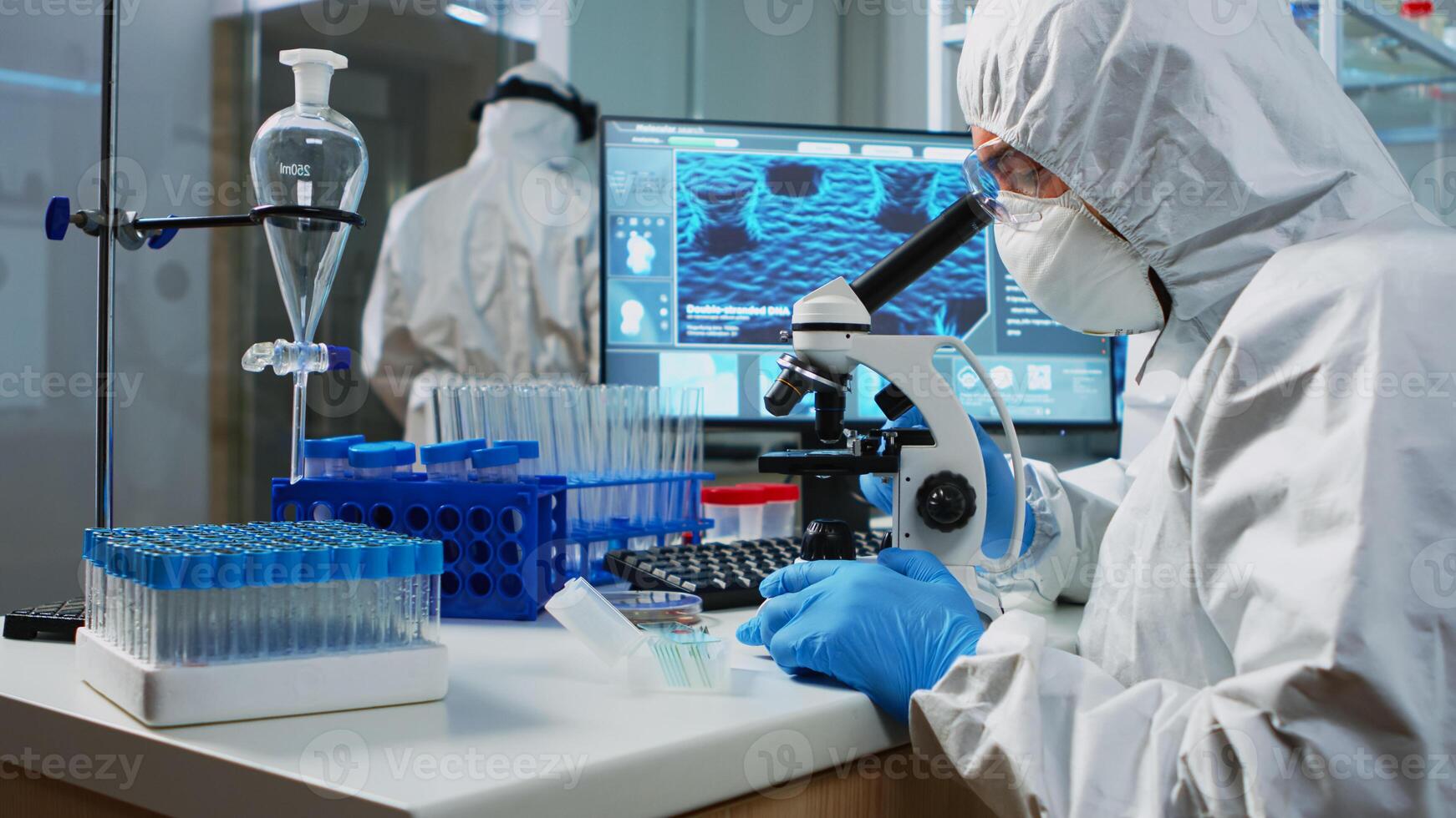 Scientist in protection suit looking through a microscope in equipped lab. Team of chemists examining virus evolution using high tech for scientific research of vaccine development against covid19. photo