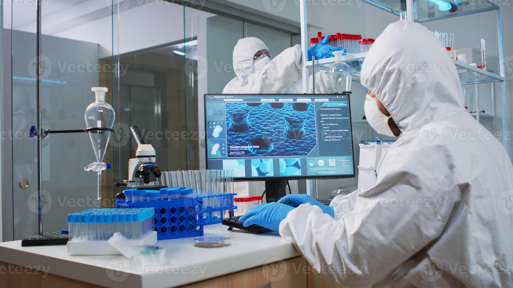 Chemist man in coverall typing on computer analysing vaccine evolution in modern equipped laboratory. Team of doctors in ppe suit using high tech for researching treatment against covid19 virus photo