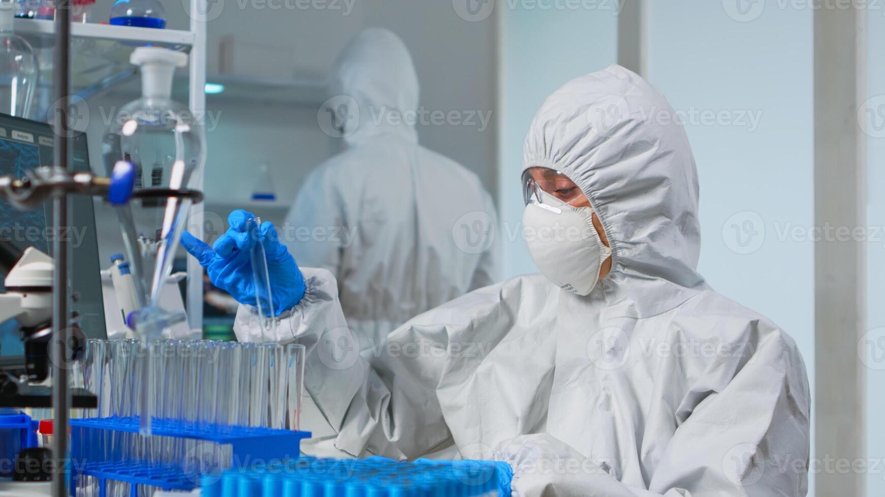 Technician in ppe suit doing experiment checking test tube in equipped lab typing results on pc. Scientist in coverall working with various bacteria tissue and blood samples for antibiotics innovation photo