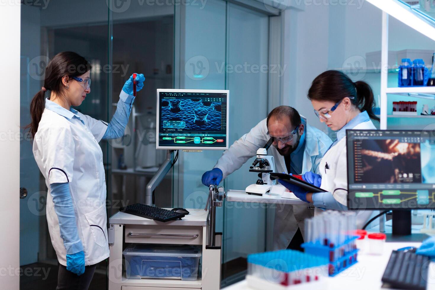 Scientist analyzing blood sample in vacutainer with researching team looking through microscope. Chemists in pharmaceutical lab examining sample for medical experiment with technology for medicine industry. photo